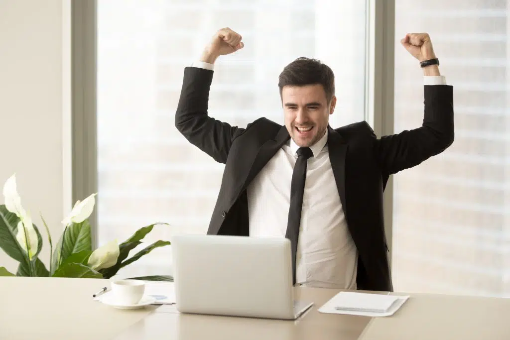 Millennial businessman raising hands and happily yelling when looking on laptop at desk. Business leader feeling positive because of successful deal confirmation, excited with great result in work